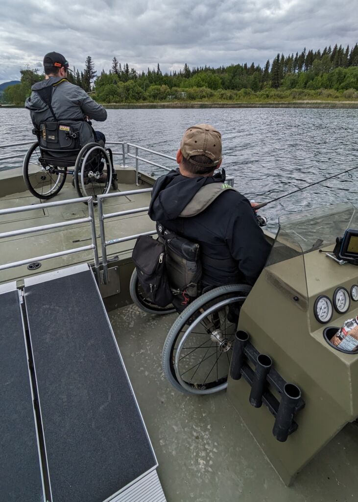 Two people sitting in wheelchairs, holding fishing rods, on two separate levels of a boat deck connected by a ramp.